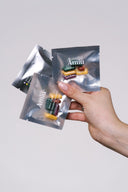 A hand holding three small silver pouches of Amíu capsules against a plain light gray background.