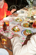 A dining table set with multiple plates of food, glasses of iced tea, and a floral table runner. A person in a bright orange top is visible on the left, and another person in a cream jacket is in the foreground, making this a lifestyle shot.