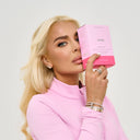 A blonde woman in a pink zip-up top holds a pink product box labeled 'Amia' next to her face against a light gray studio background.
