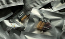 Several silver Amíu Daily Stack single-serve packets are arranged on a dark surface, with one packet in front showing capsules inside.