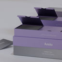 A stack of Amiu branded product boxes in lavender and gray packaging, with one box open to reveal a dark interior tray, photographed on a clean light gray background.