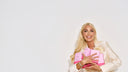 A blonde woman in a cream satin blazer holds several bright pink product boxes against a clean white studio background.
